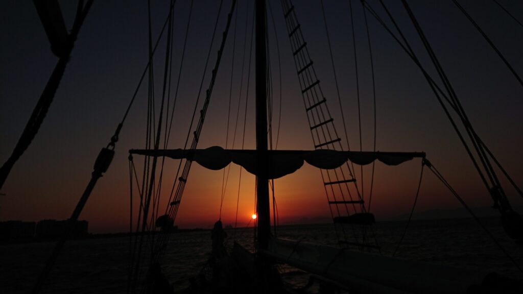 Silhouetted ship mast, sail, and rigging against a glowing orange sunset over the sea.